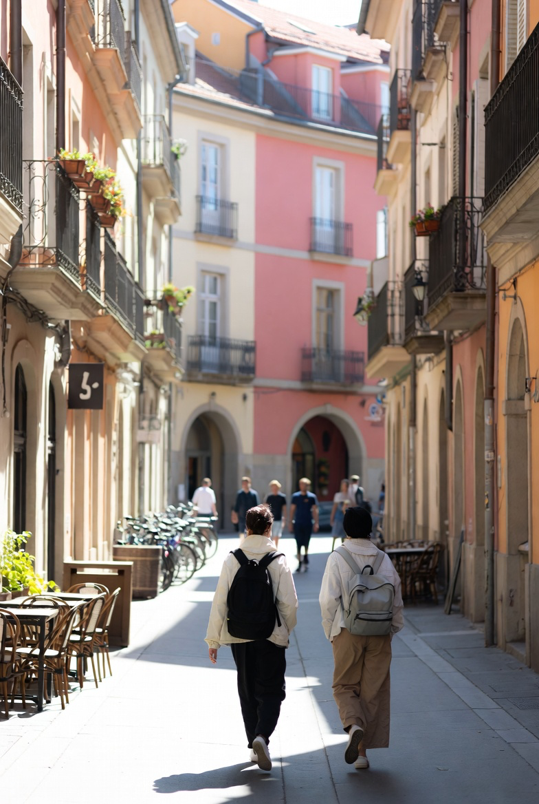 Two friends walking down a sunlit cobblestone European old-town street on a weekend morning